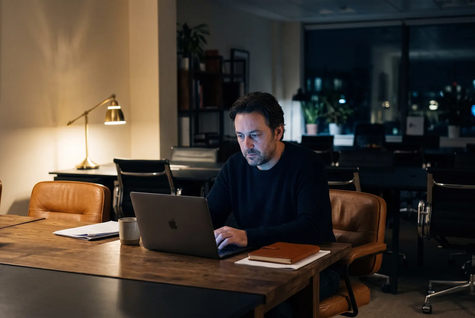 A man works alone at a laptop late at night in a dark office