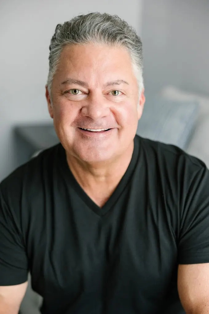 John Nichols, smiling, seated in his office in a dark V-neck shirt